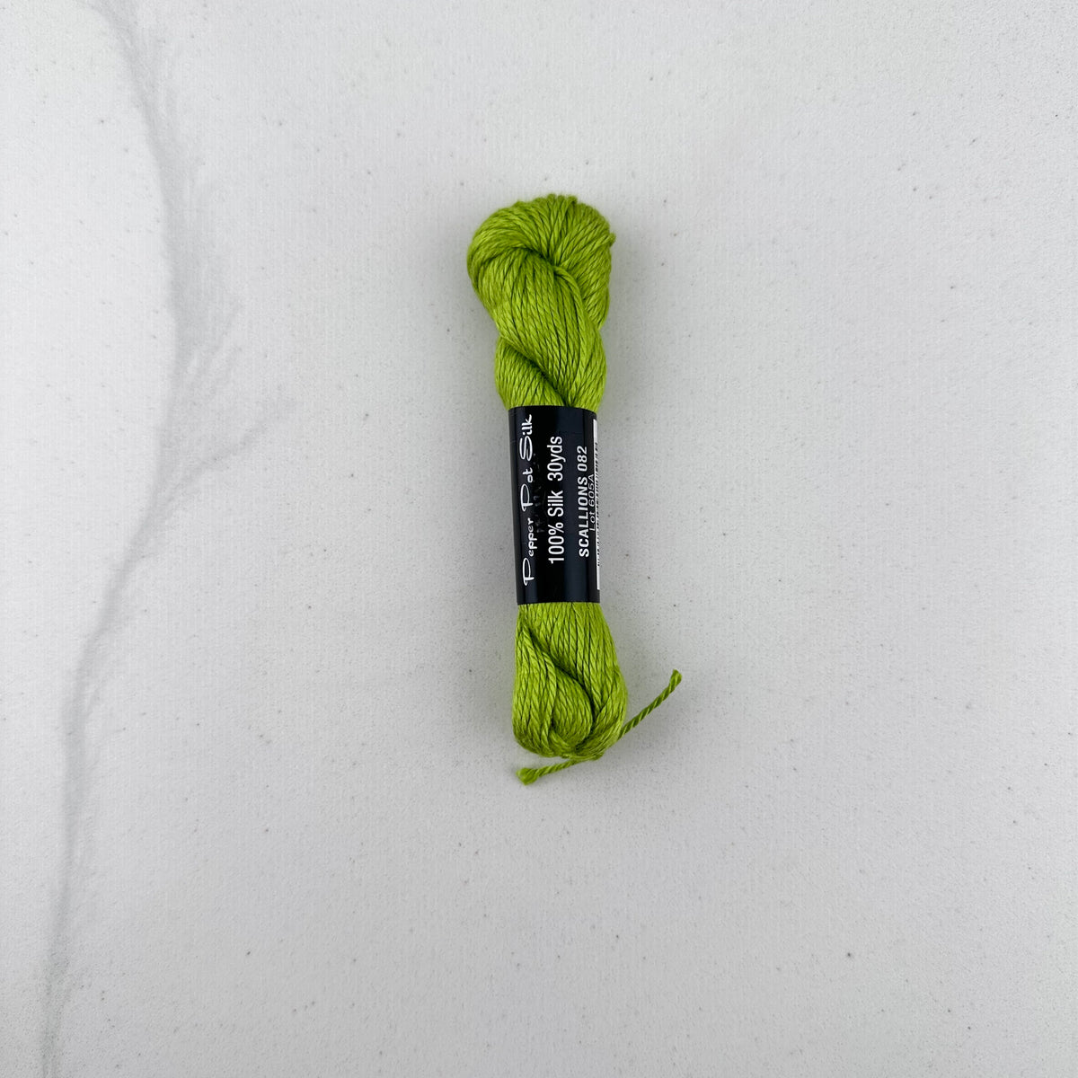 30 yard skein of Pepper Pot Silk needlepoint thread in a green scallion 082, with black and white label lying flat on white background