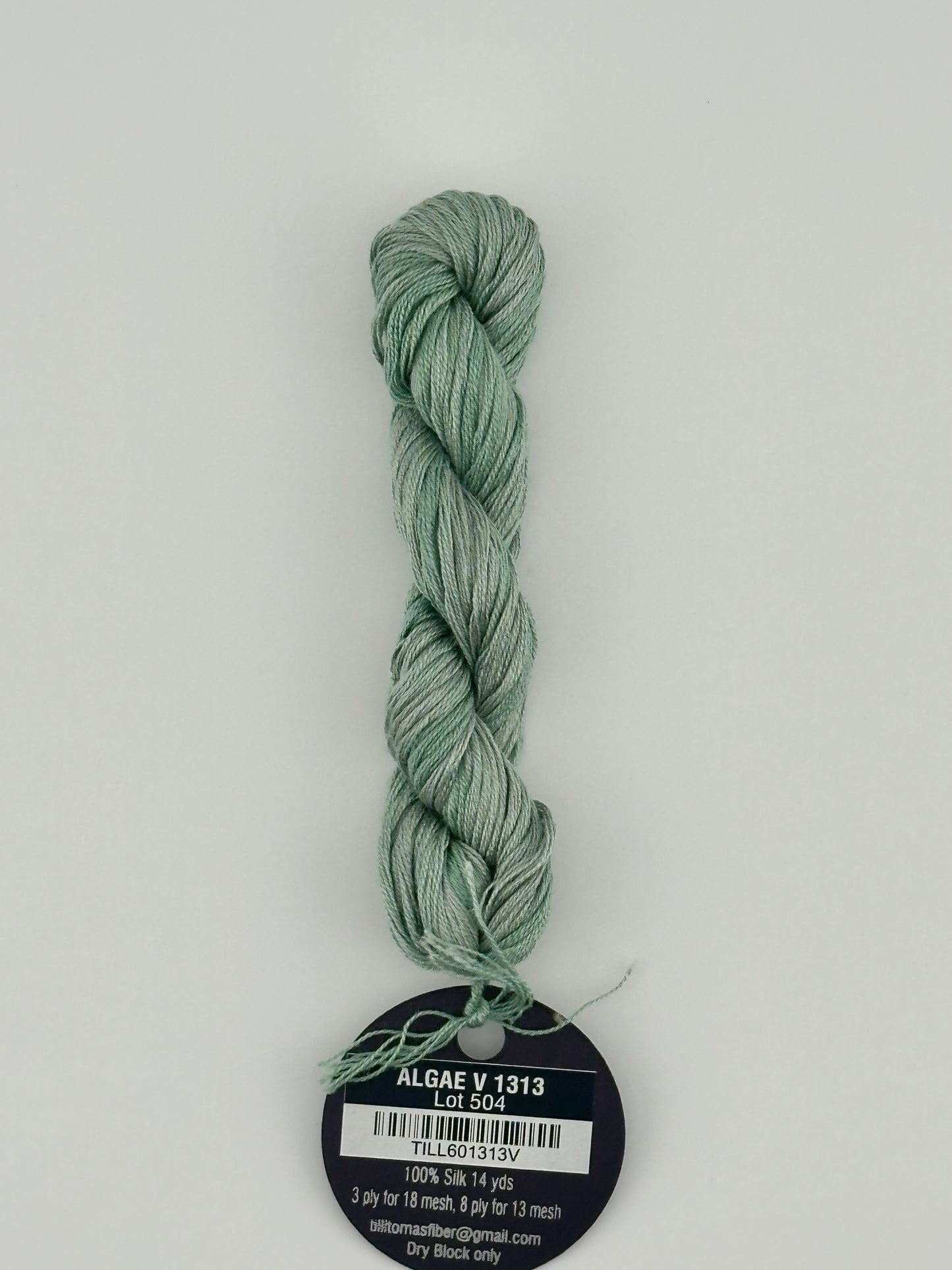 14 yard skein Planet Earth Silk 4 Ply needlepoint thread, in algae v1313 green, with round black and white label lying flat on white background.