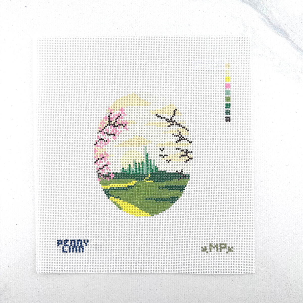 18 mesh needlepoint canvas featuring a circular design ox the emerald city with green fields, pink cherry blossoms, a pale yellow sky, and dark branch silhouettes. The canvas has the text 'PENNY LINN on the bottom left and a small vertical color swatch on the right side. laying flat on white background.