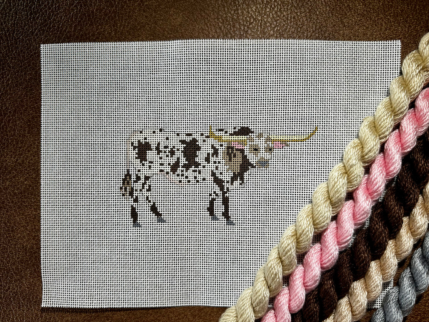A needlepoint canvas featuring a brown-and-white bull, placed on a brown leather surface with thread skeins in beige, pink, brown, and gray diagonally crossing the lower right corner.