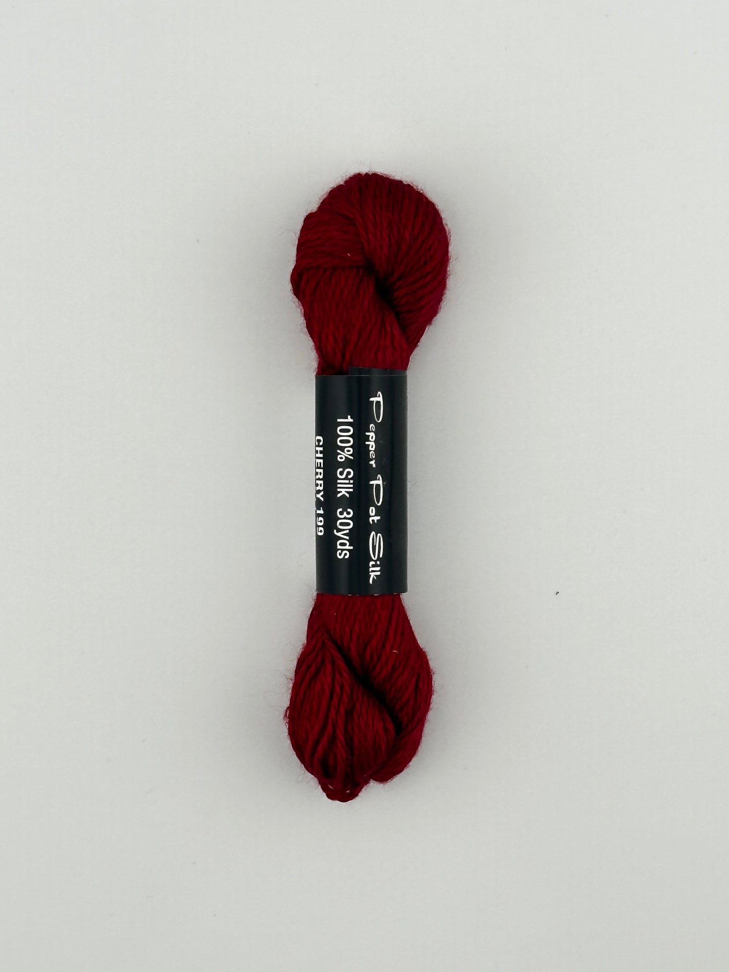 30 yard skein of Pepper Pot Silk needlepoint thread in cherry 199, with black and white label lying flat on white background