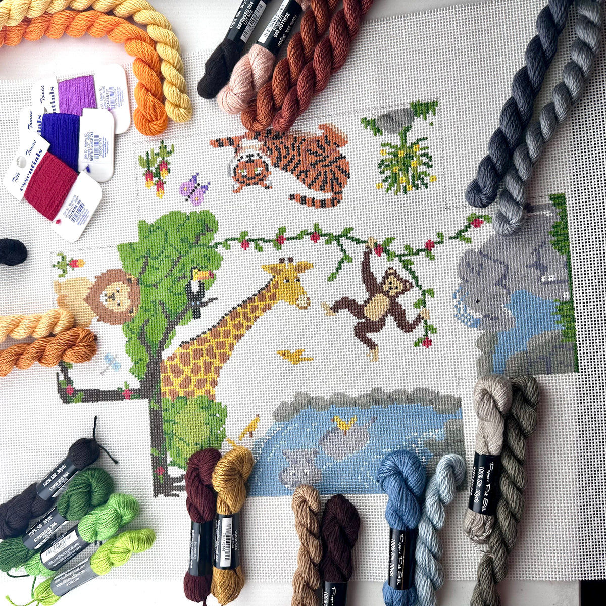 13 mesh needlepoint brick cover canvas featuring a colorful safari animal design on a white fabric background. The design includes a lion, giraffe, monkey, tiger, elephant, and a hippo in a pond.
Shown with threads in matching colors.