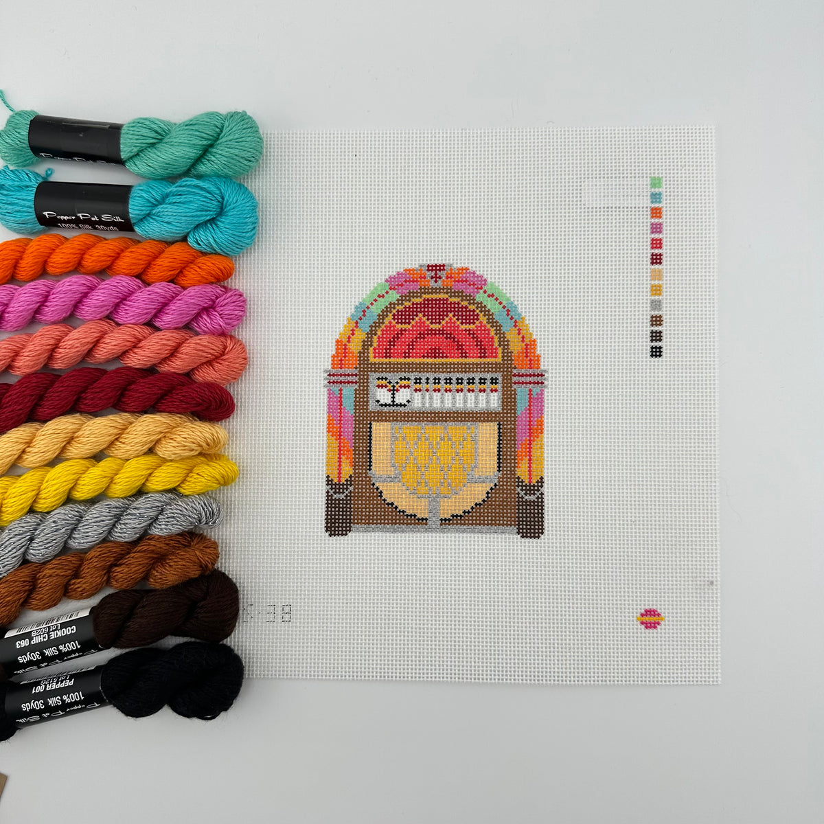 A needlepoint canvas featuring a vintage jukebox design by saturnalia kitted with12 skeins of thread to match the color key. canvas and threads lying flat against a plain white background.