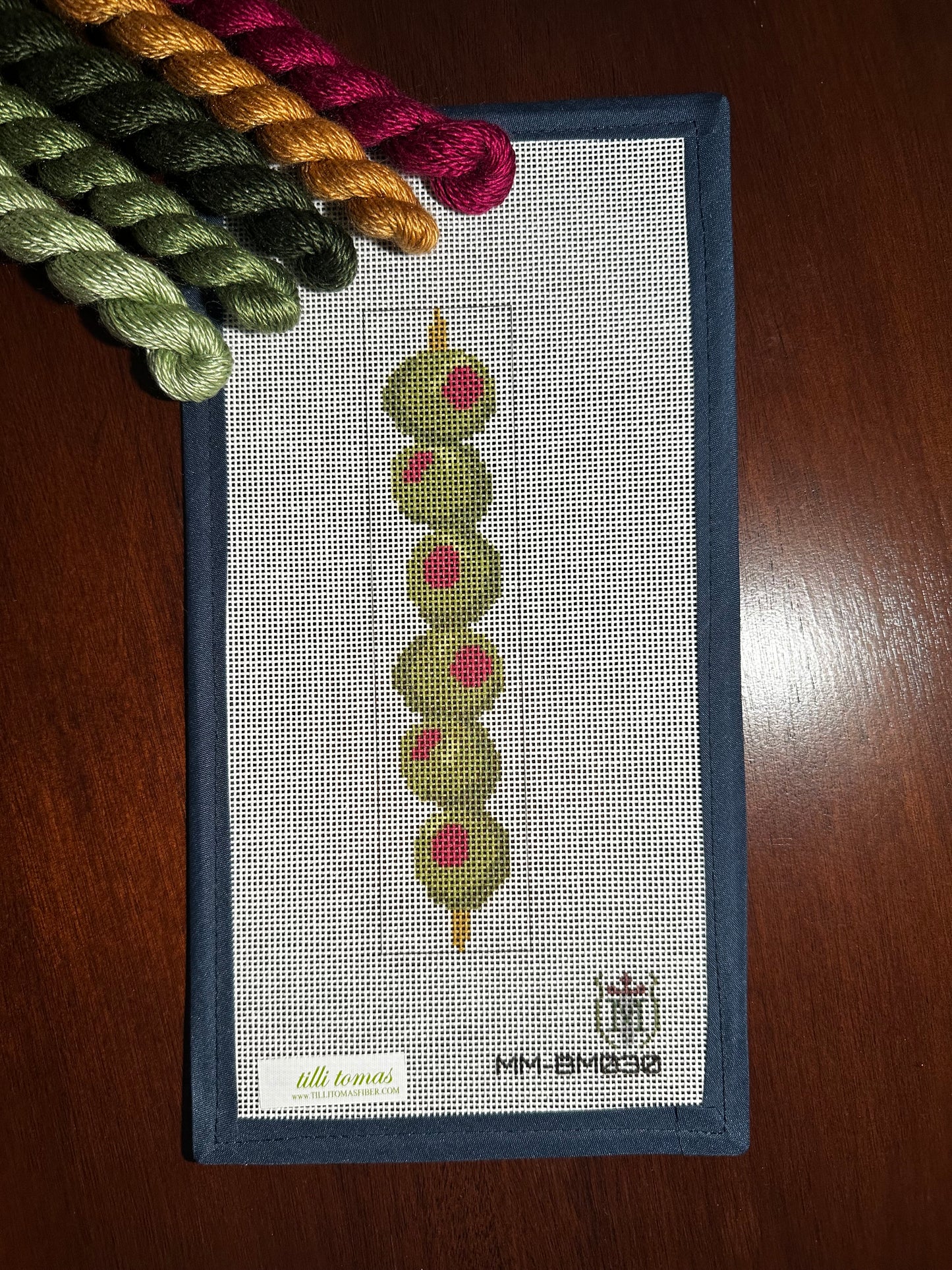 A needlepoint canvas featuring a vertical string of green olives with pink centers, bordered in dark blue. The canvas is displayed on a dark wooden surface with matching thread skeins placed in the top-left corner.