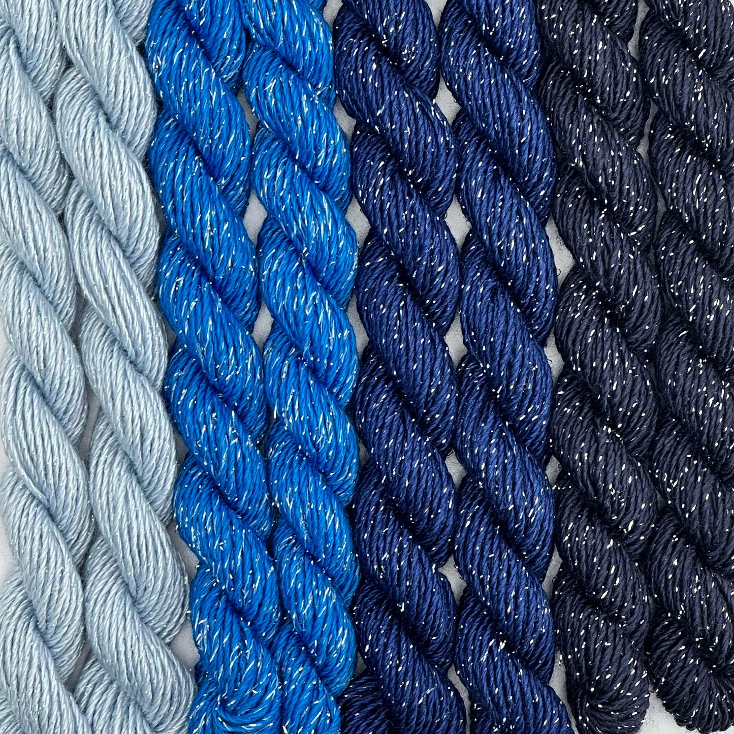 close-up of 8 skeins of Stardust Silk and Ivory needlepoint thread in four shades of blue including blue yonder 606, midnight 531, blue streak 592, and regatta 736, lying side by side.