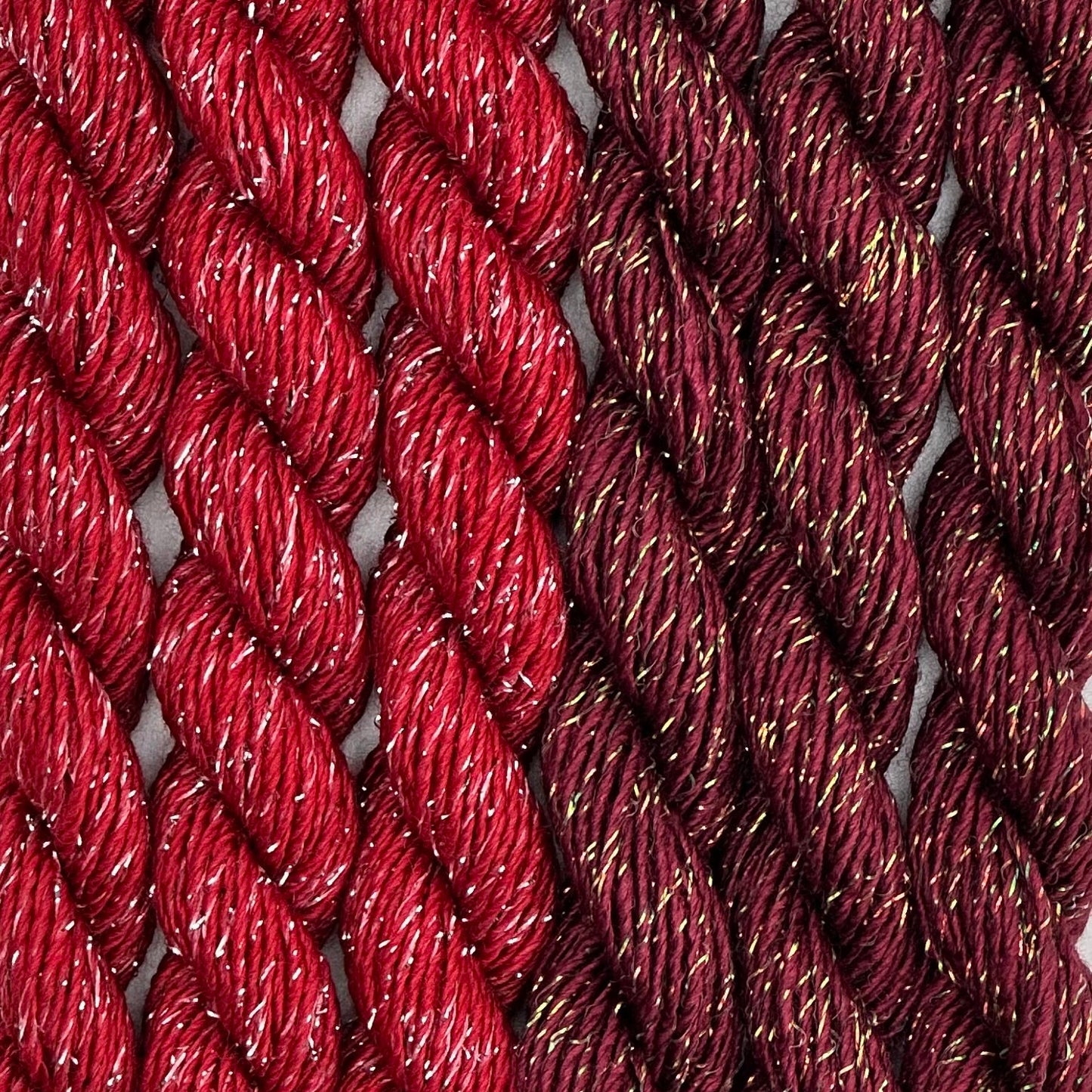 close-up of 6 skeins of Stardust Silk and Ivory needlepoint thread in bordeaux 543 and ramblin rose 664 reds, lying side by side.