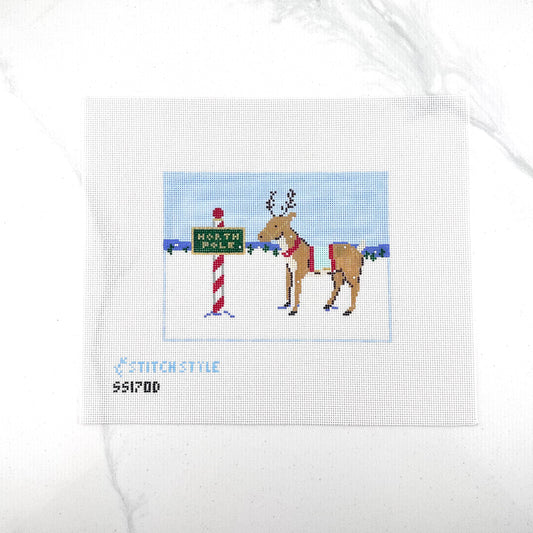 18 mesh needlepoint canvas featuring a Christmas scene with a brown reindeer wearing a red harness, standing next to a red-and-white striped pole with a green sign that reads 'NORTH POLE'. With the brand name 'STITCHSTYLE' and number 'S51700' printed in blue at the bottom left.