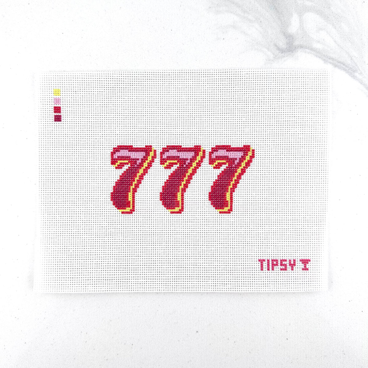 A close-up of a needlepoint canvas showing the number 777 stitched in dark red and pink with a yellow outline, on a white 18 mesh grid. The brand name 'TIPSY' is visible in the bottom right corner.