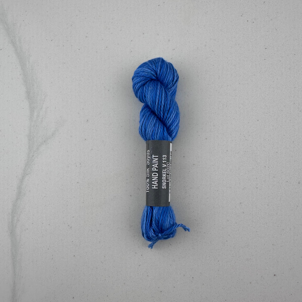30 yard skein of Pepper Pot Silk needlepoint thread in a blue variegated snorkel v113, with black and white label lying flat on white background