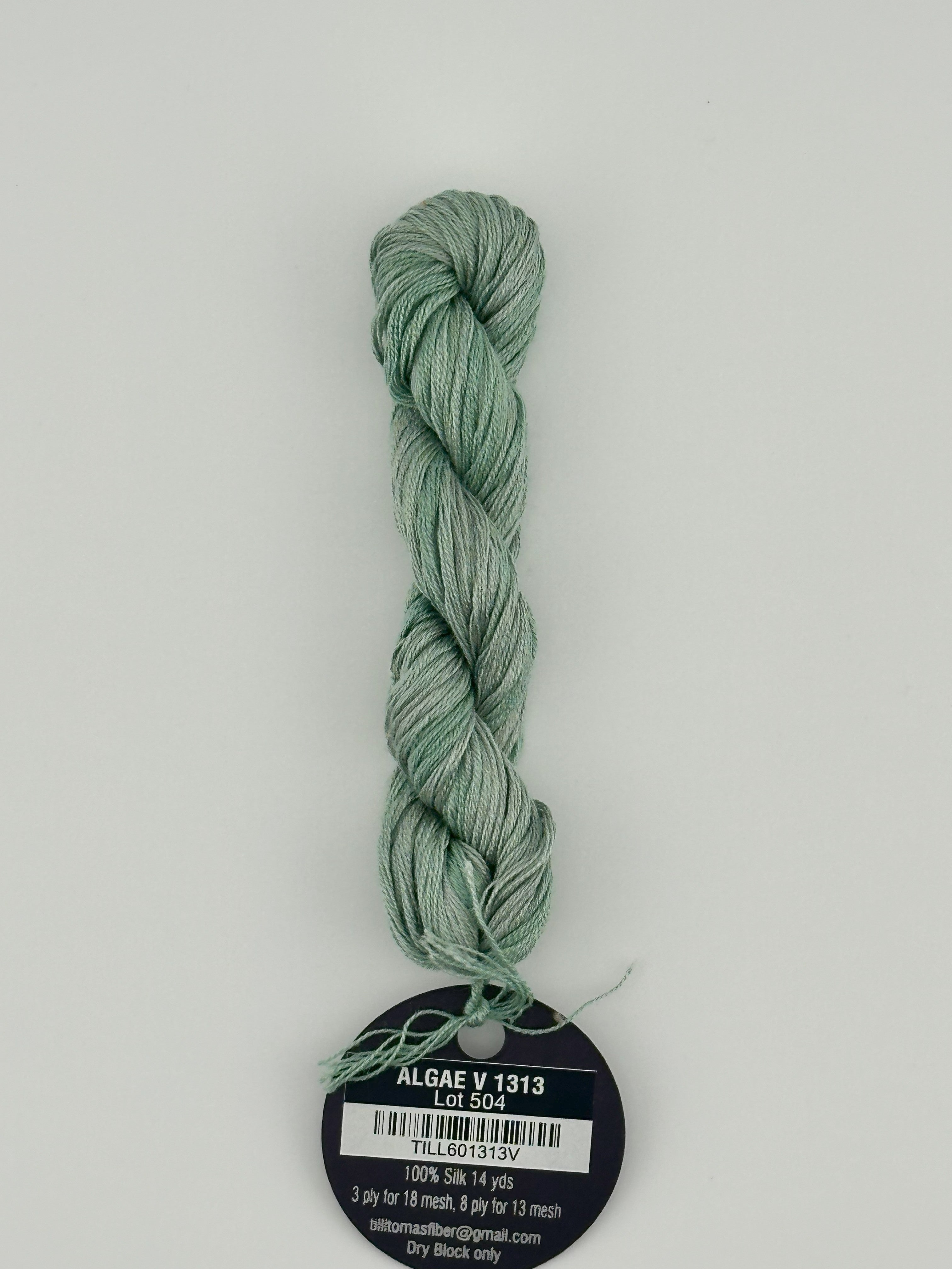 14 yard skein Planet Earth Silk 4 Ply needlepoint thread, in algae v1313 green, with round black and white label lying flat on white background.