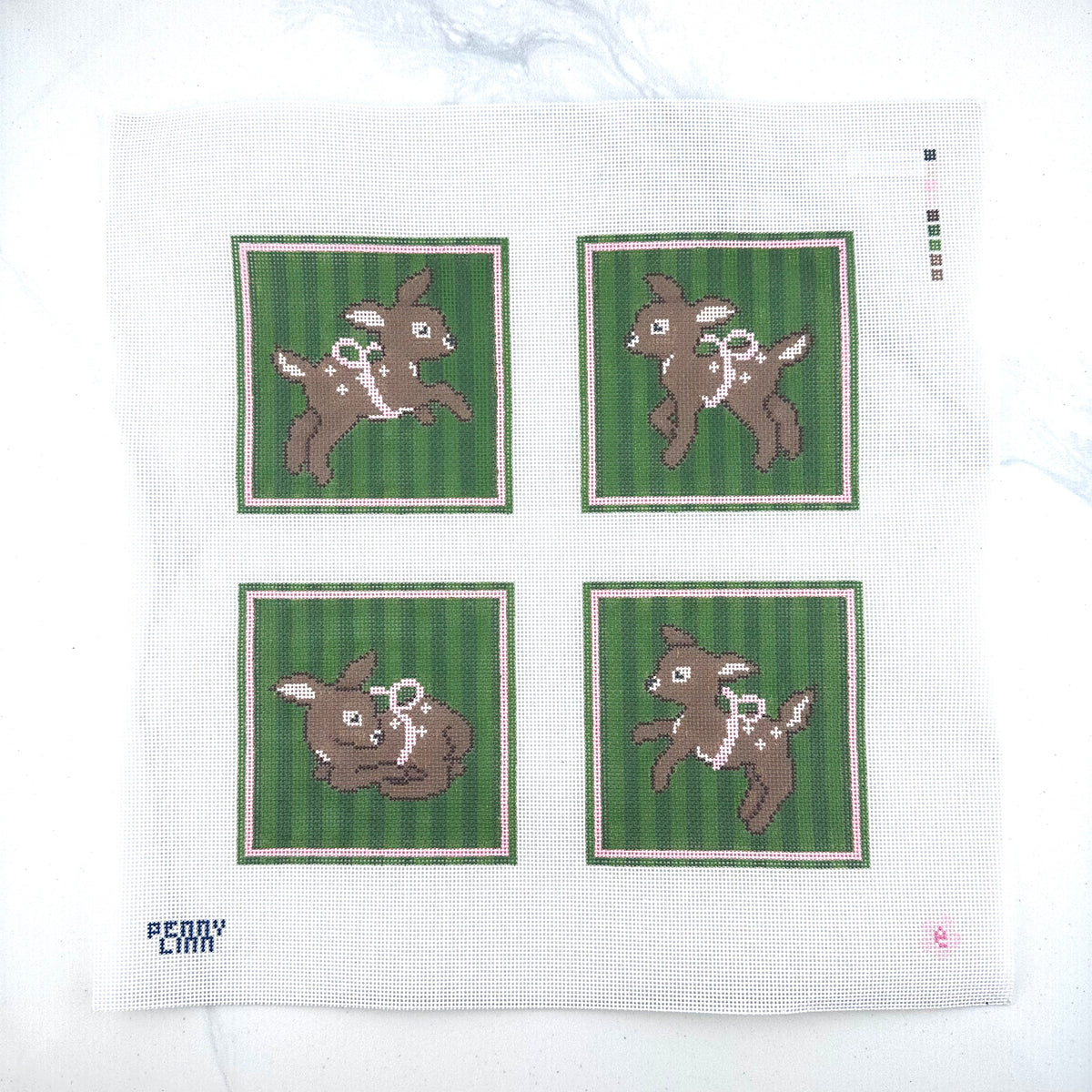 A needlepoint canvas featuring four square coasters with brown fawns on green striped backgrounds, placed on a white surface. The brand name 'PENNY LINN" is visible in the bottom-left corner.