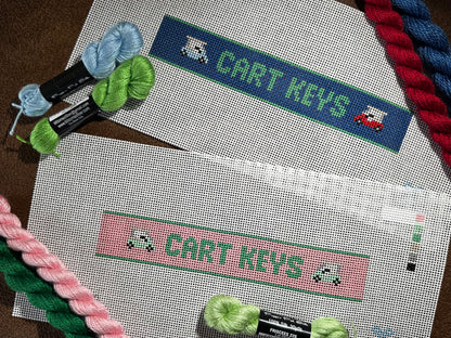 Two needlepoint key fob canvas featuring a pink and green sign  and blue and green sign with the text 'CART KEYS' in green on 14 mesh lying flat on brown background with matching colored thread skeins laid out on top.
