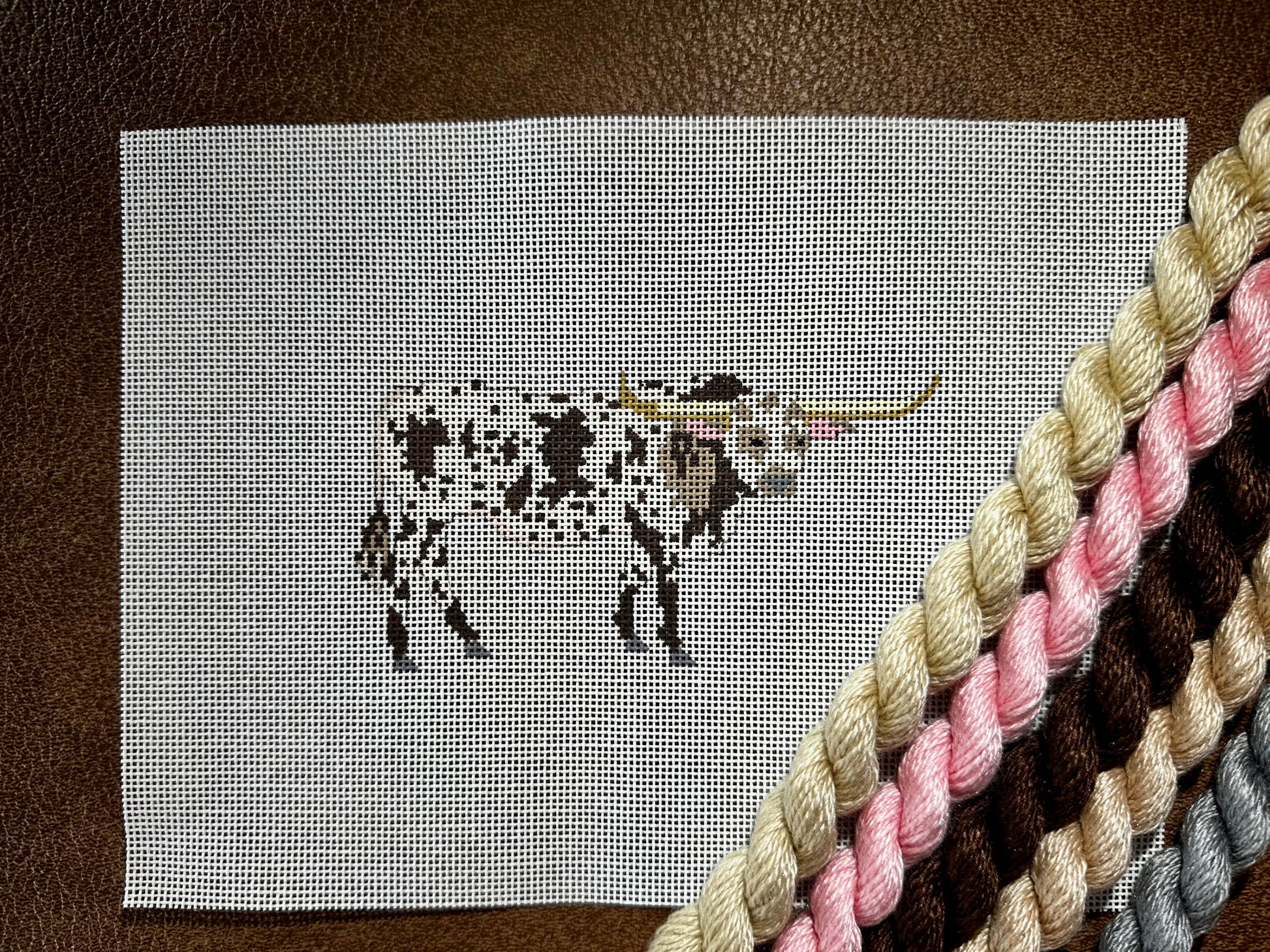 A needlepoint canvas featuring a brown-and-white bull, placed on a brown leather surface with thread skeins in beige, pink, brown, and gray diagonally crossing the lower right corner.