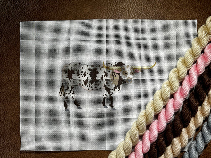 A needlepoint canvas featuring a brown-and-white bull, placed on a brown leather surface with thread skeins in beige, pink, brown, and gray diagonally crossing the lower right corner.