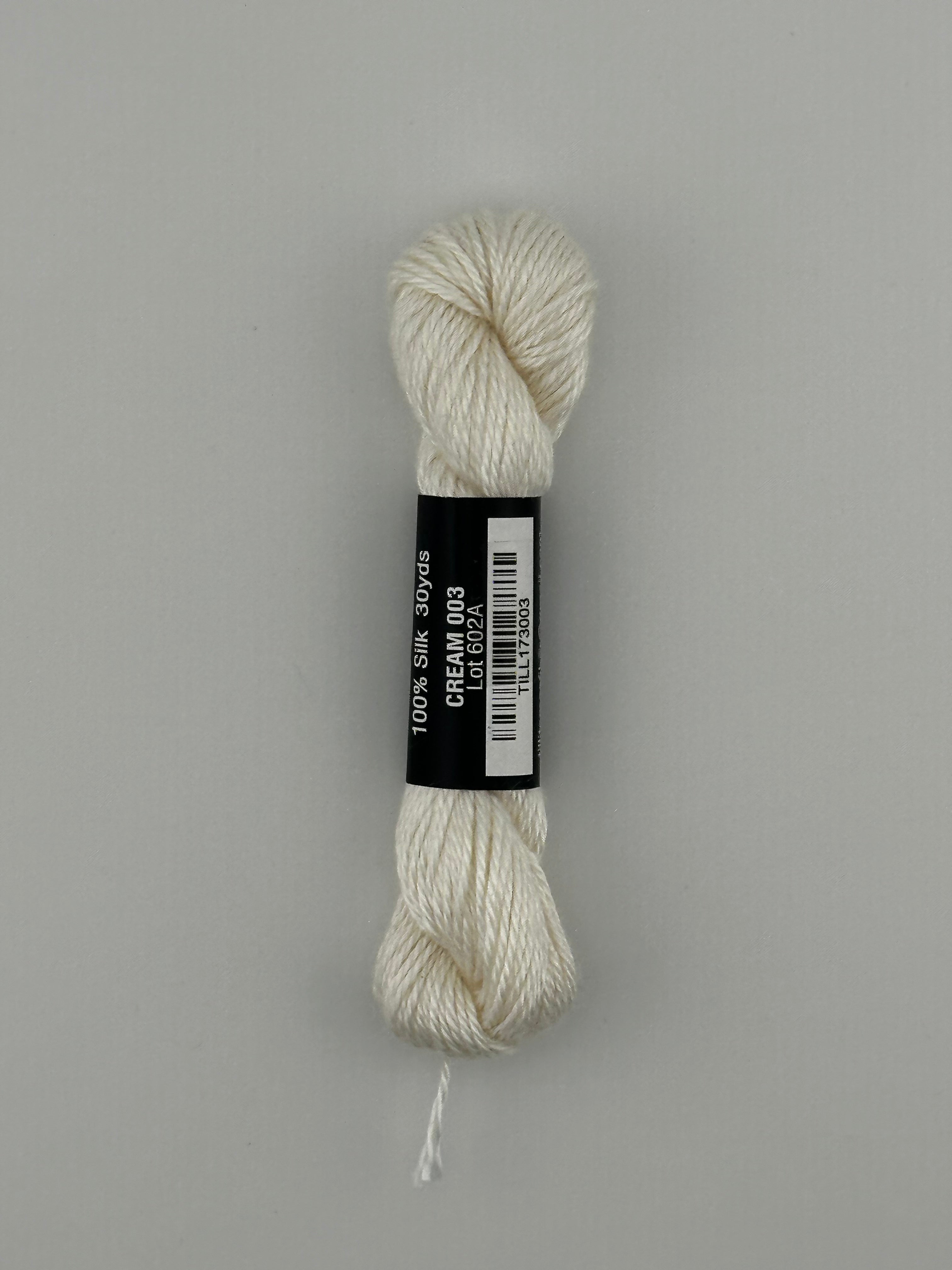 30 yard skein of Pepper Pot Silk needlepoint thread in cream 003, with black and white label lying flat on white background