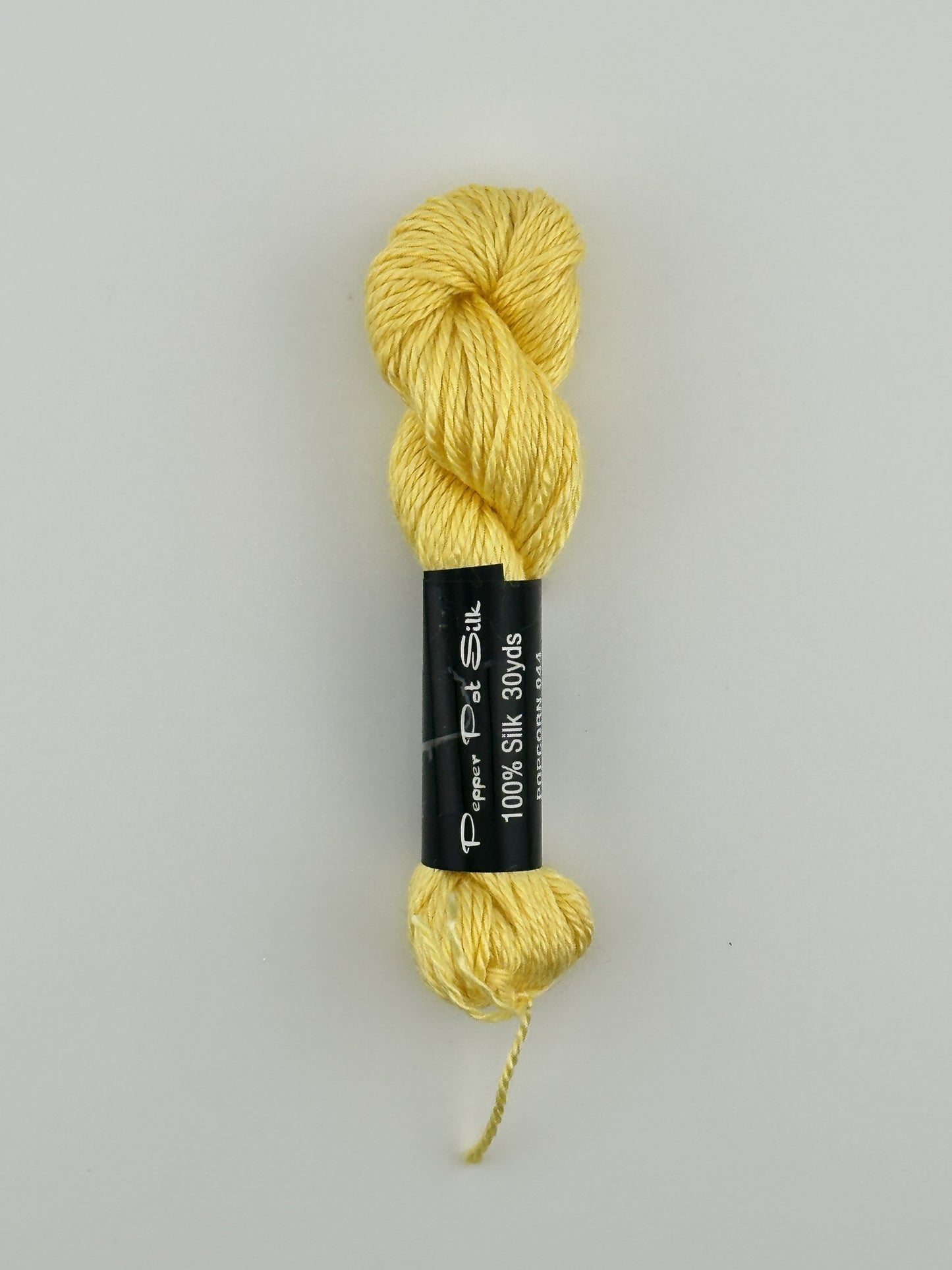 30 yard skein of Pepper Pot Silk needlepoint thread in popcorn 044 yellow, with black and white label lying flat on white background