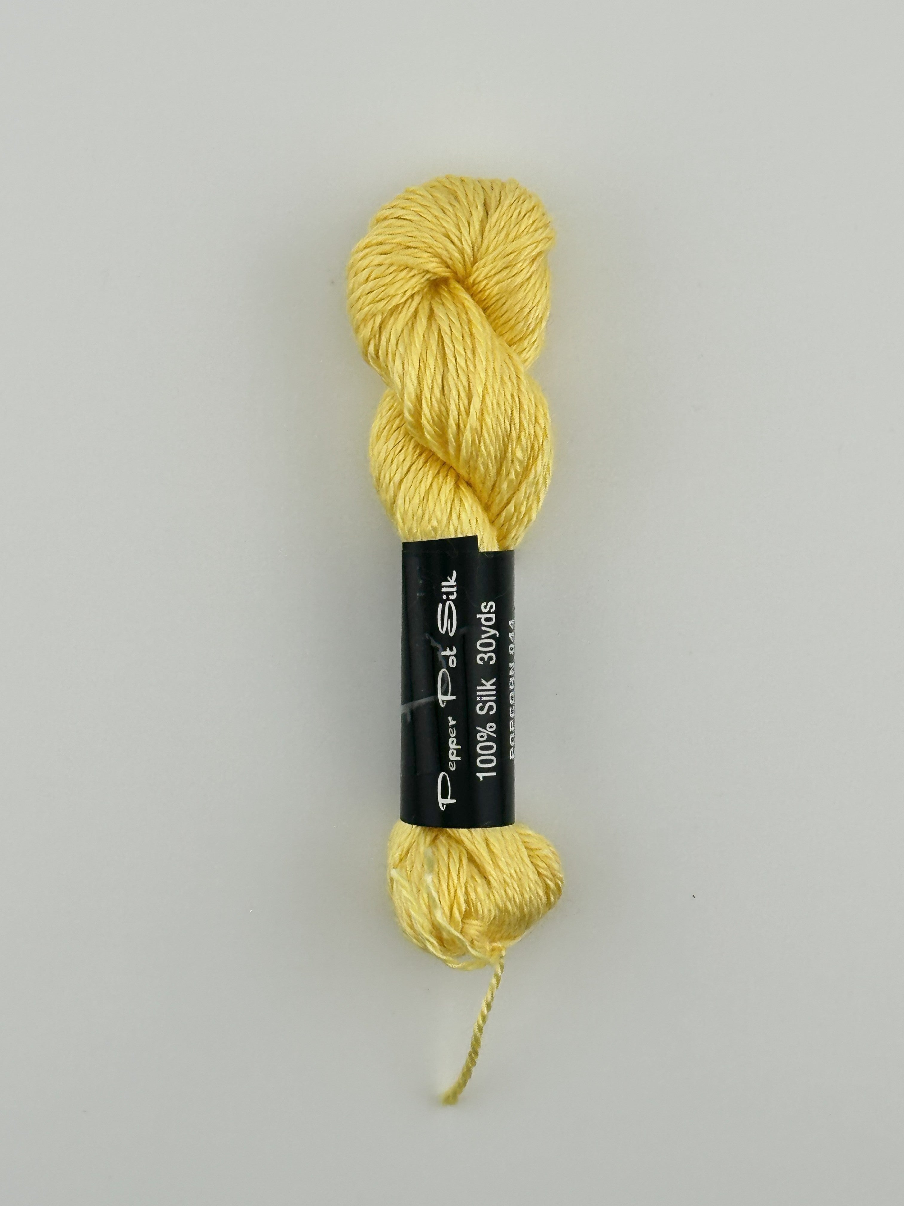 30 yard skein of Pepper Pot Silk needlepoint thread in popcorn 044 yellow, with black and white label lying flat on white background