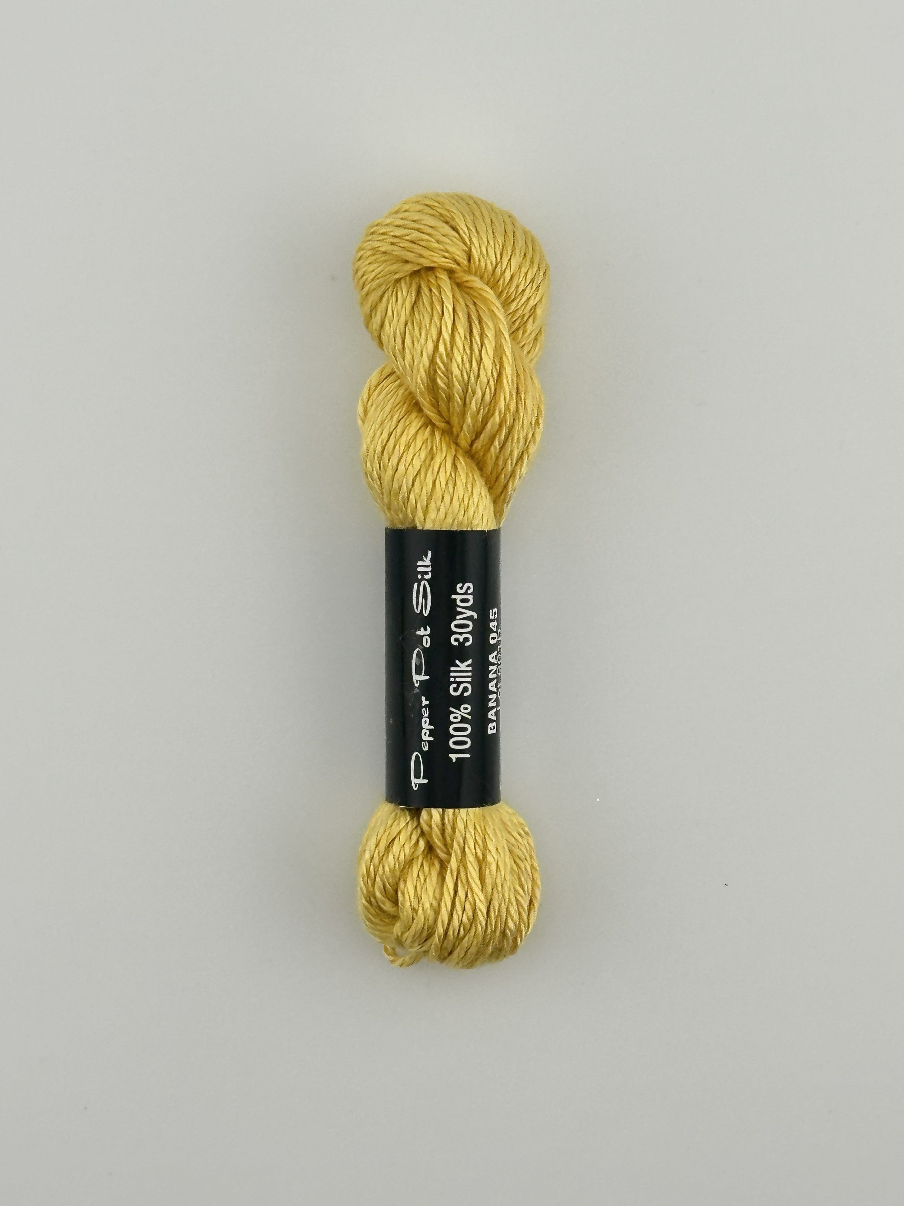 30 yard skein of Pepper Pot Silk needlepoint thread in banana 045 yellow, with black and white label lying flat on white background