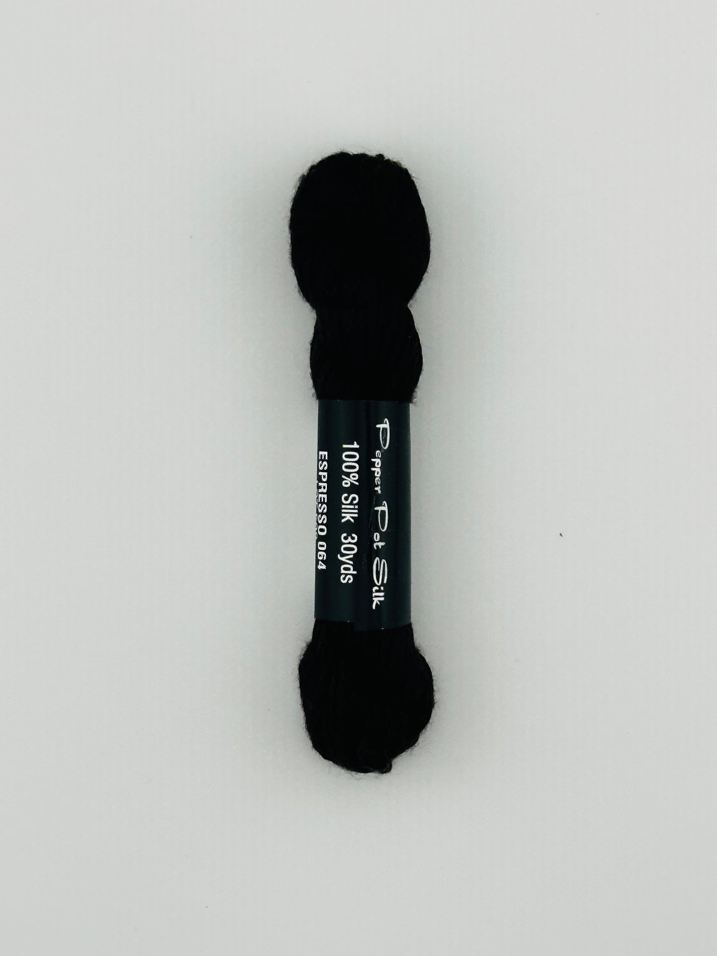 30 yard skein of Pepper Pot Silk needlepoint thread in espresso 064, with black and white label lying flat on white background