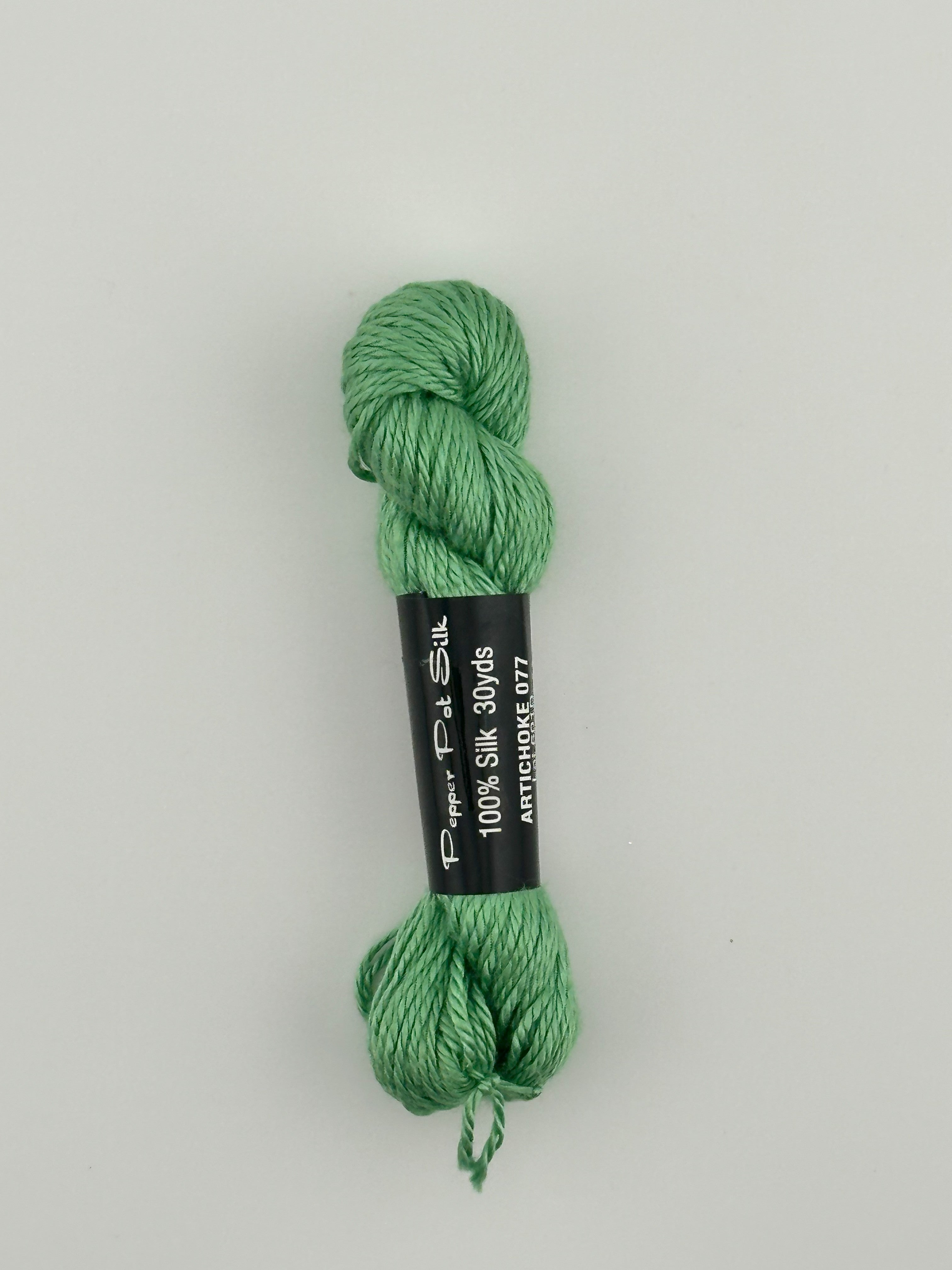 30 yard skein of Pepper Pot Silk needlepoint thread in Artichoke 077 green, with black and white label lying flat on white background