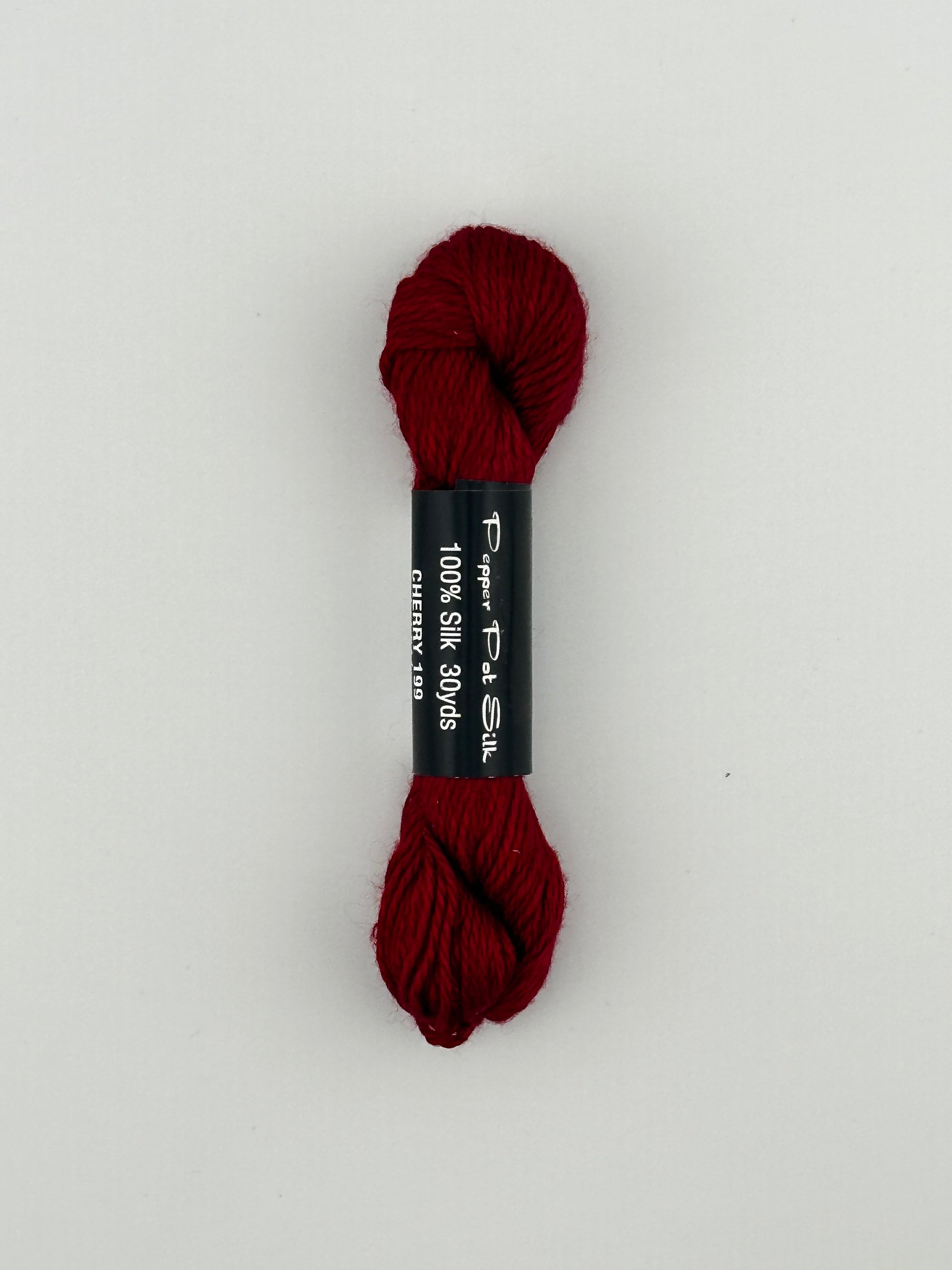 30 yard skein of Pepper Pot Silk needlepoint thread in cherry 199, with black and white label lying flat on white background
