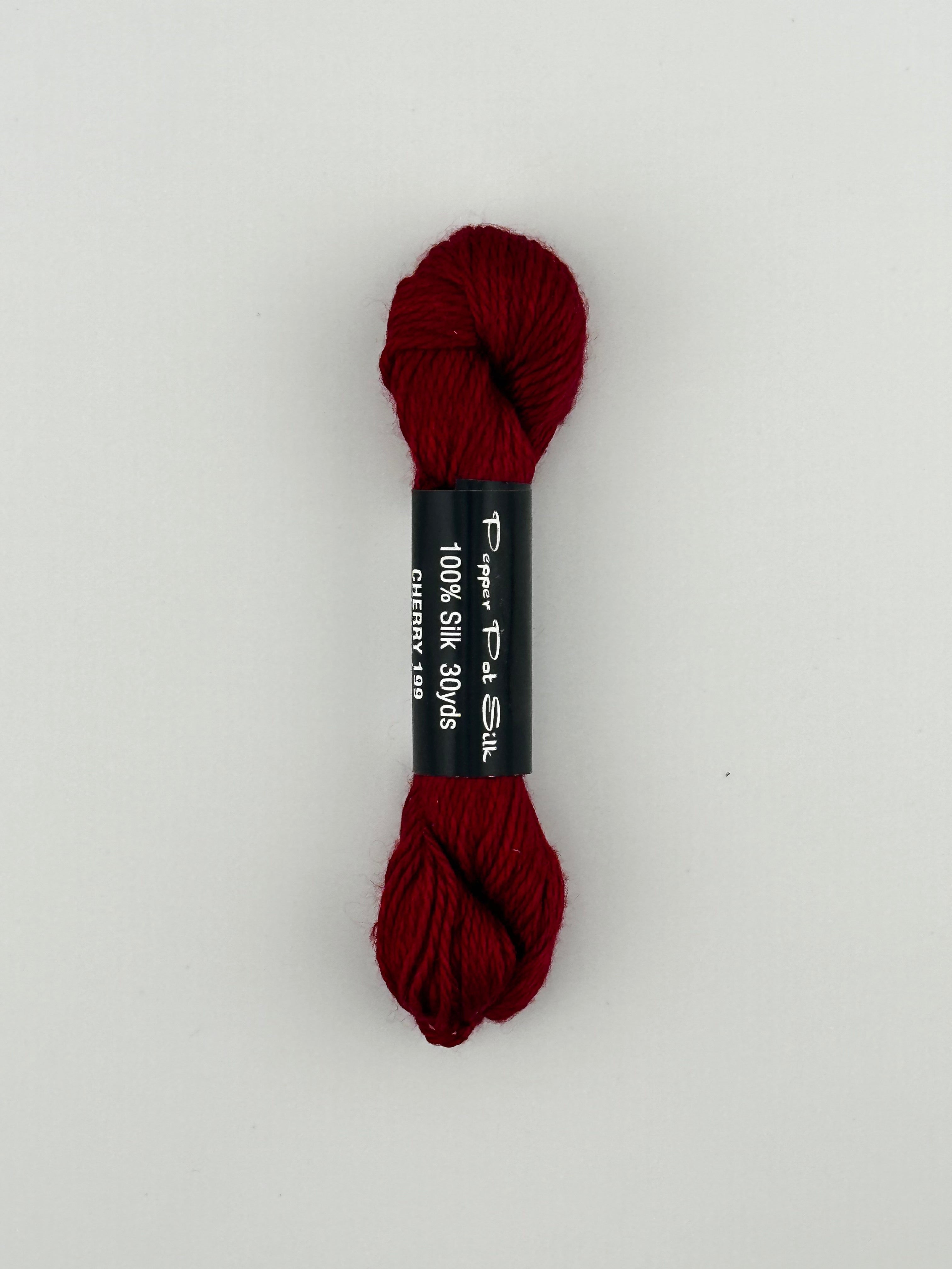 30 yard skein of Pepper Pot Silk needlepoint thread in cherry 199, with black and white label lying flat on white background