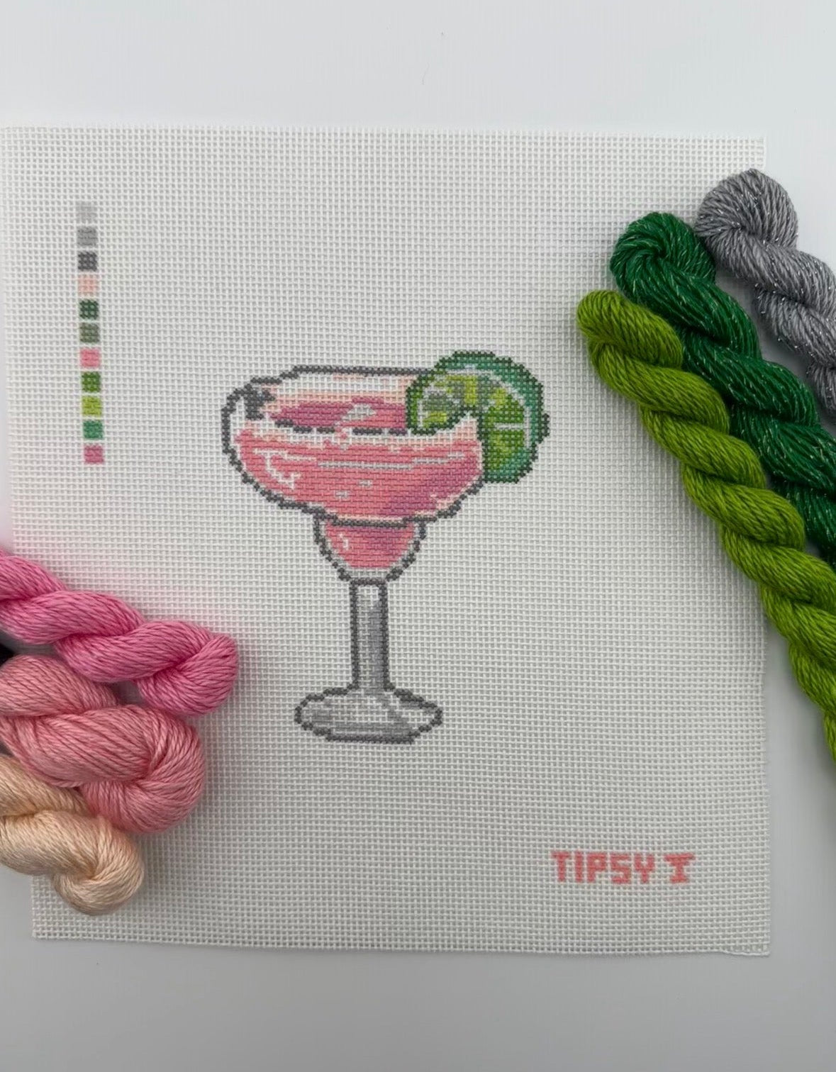 A square 18 mesh needlepoint canvas featuring a pink margarita cocktail in a glass with a lime wedge, on a white background with a small red TIPSY  text at the bottom right and matching threads on top of canvas.