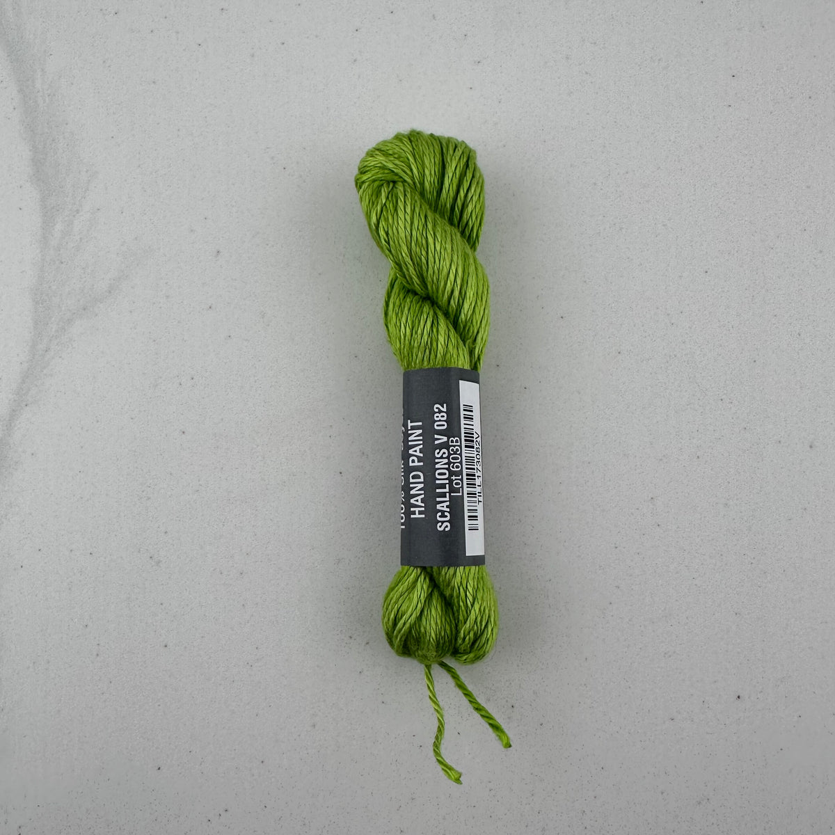 30 yard skein of Pepper Pot Silk needlepoint thread in a green variegated scalllion v082, with black and white label lying flat on white background