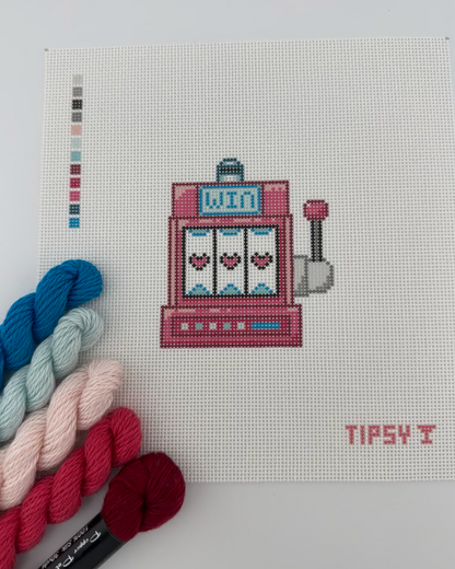 18 mesh needlepoint canvas with pink slot machine with three hearts on the reels, a small blue sign reading 'WIN', and a lever on the right. The canvas is photographed against a white background, with a small color swatch strip on the left and the text TIPSY in the lower right corner. And pink and blue threads matching the color swatch laid next to canvas.