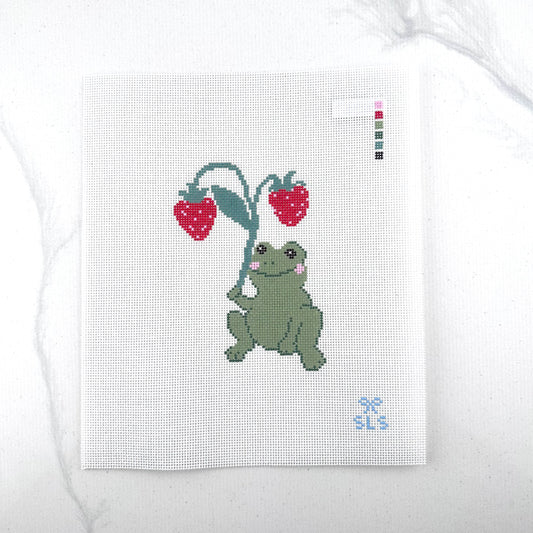 an 18 mesh needlepoint canvas featuring a design of a green frog holding a leafy plant with two red strawberries, photographed on a white background.