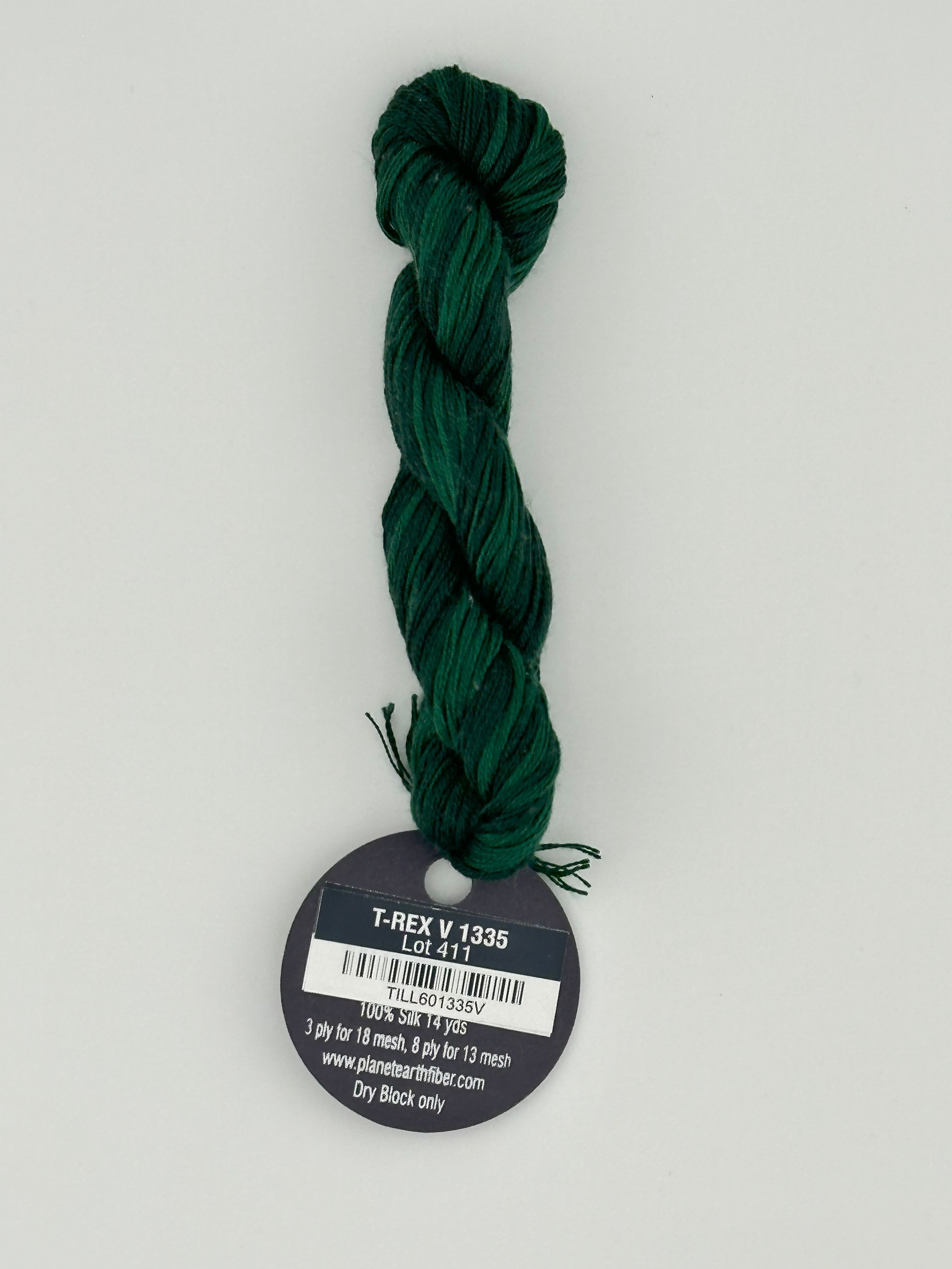 14 yard skein Planet Earth Silk 4 Ply needlepoint thread, in t-rex v 1335  green with round black and white label lying flat on white background.