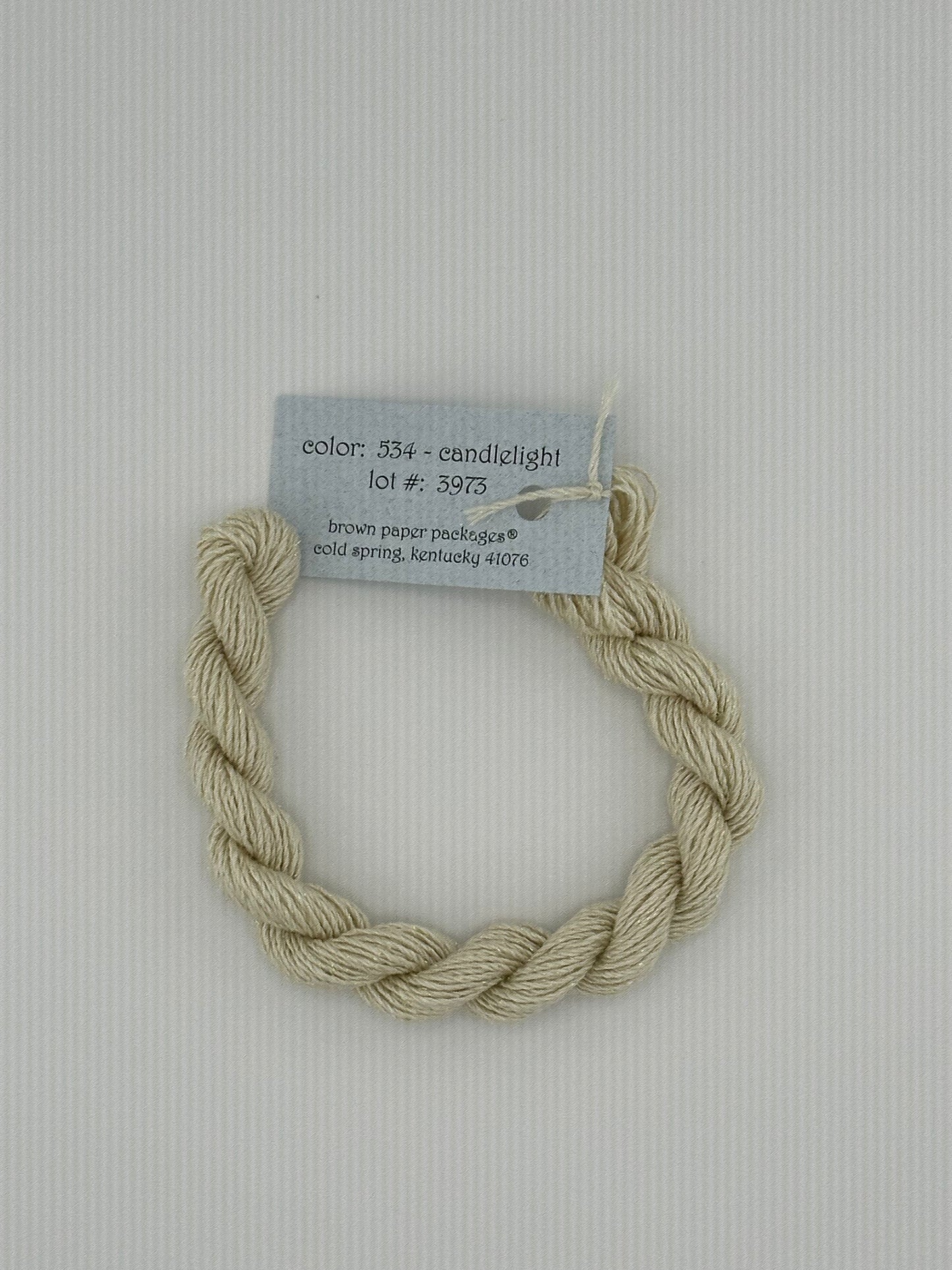 30 yard skein of Stardust Silk and Ivory needlepoint thread in Candlelight 534, with light blue label lying flat on white background