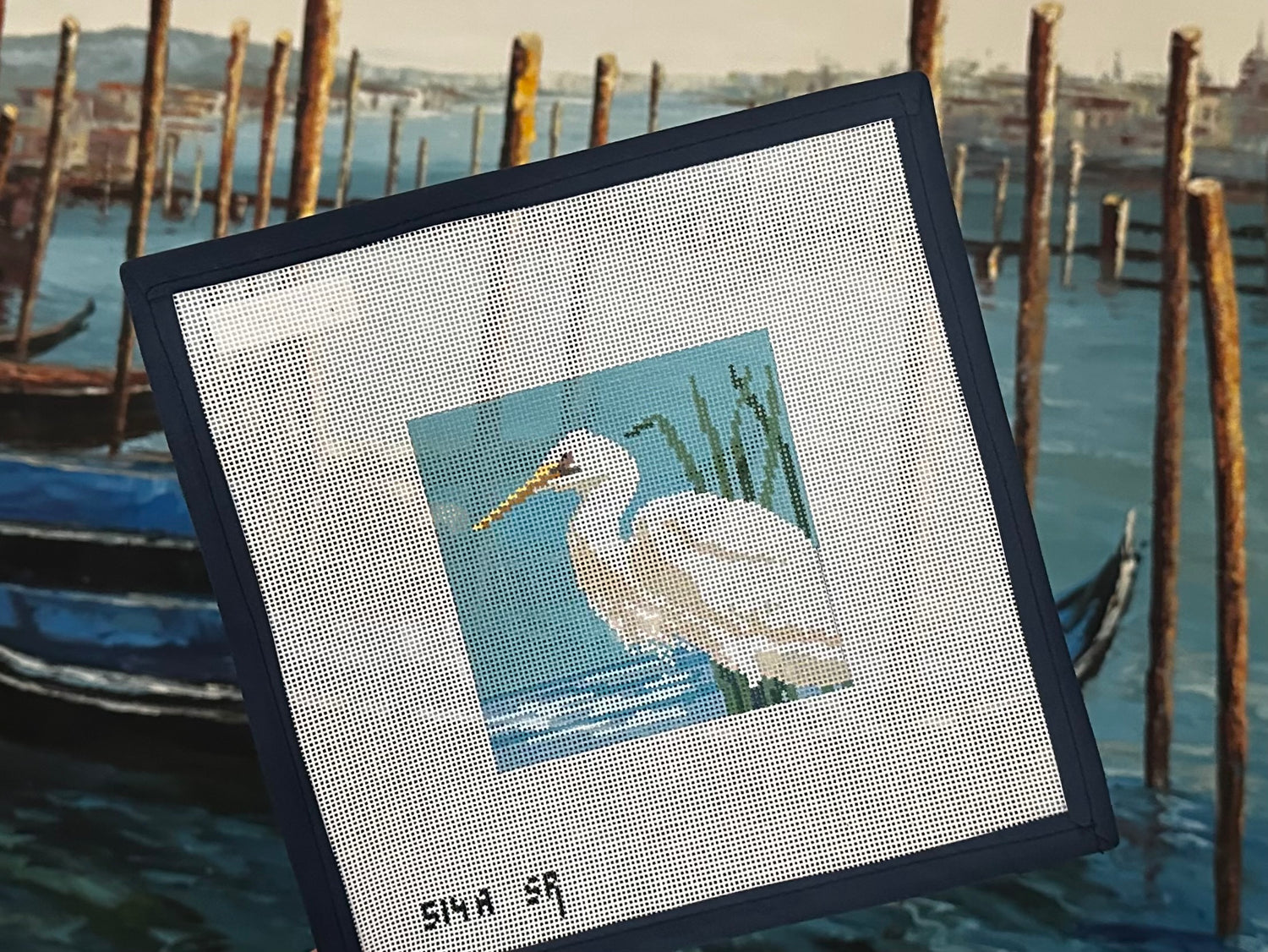 A needlepoint canvas featuring a white egret in a blue marsh, shown at an angle against a blurred background of a Venice canal with gondolas and wooden posts.