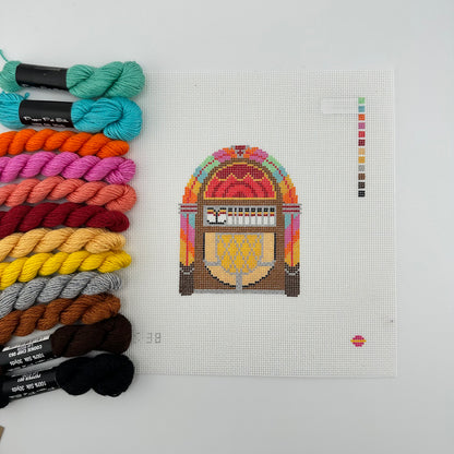 A needlepoint canvas featuring a vintage jukebox design by saturnalia kitted with12 skeins of thread to match the color key. canvas and threads lying flat against a plain white background.