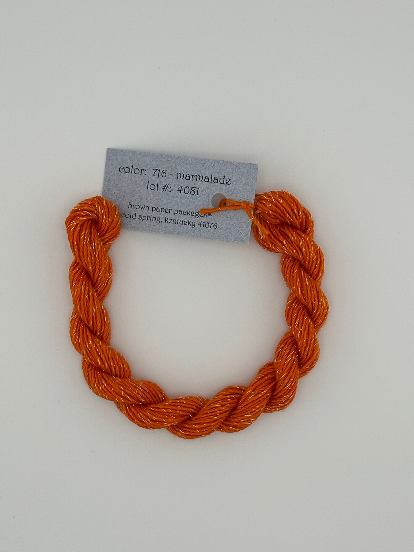 30 yard skein of Stardust Silk and Ivory needlepoint thread in Marmalade 716 orange, with light blue label lying flat on white background