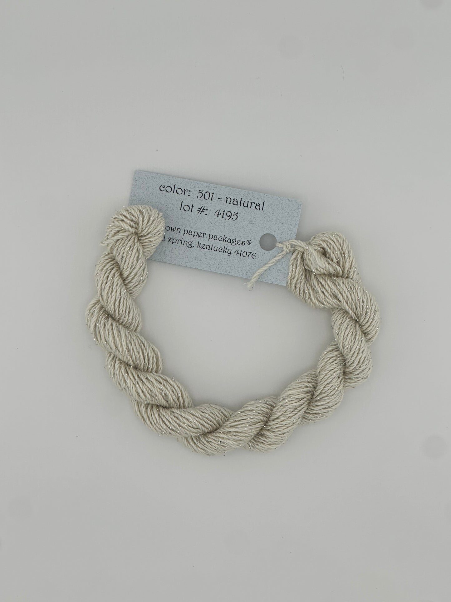 30 yard skein of Stardust Silk and Ivory needlepoint thread in Natural 501, with light blue label lying flat on white background