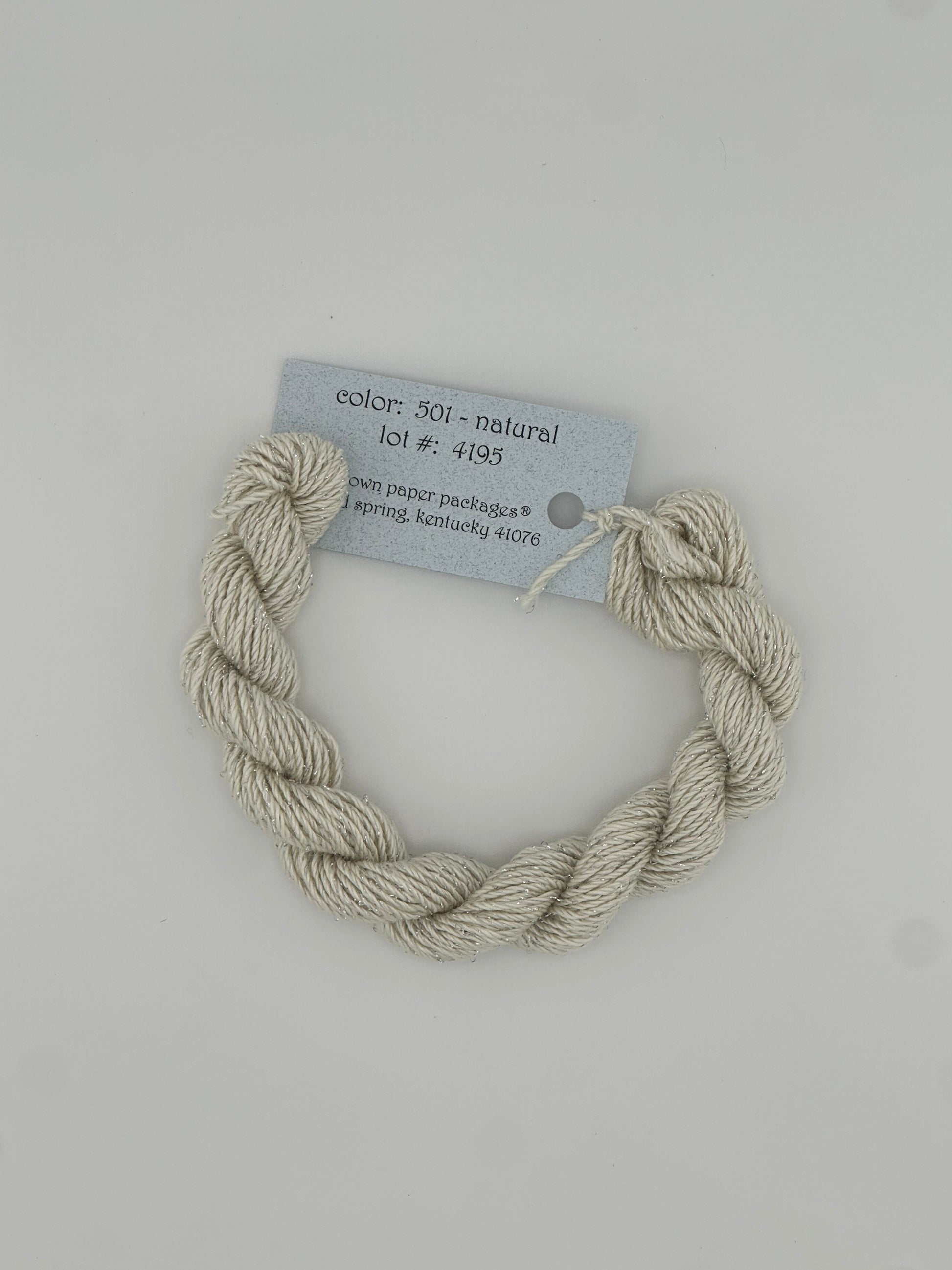 30 yard skein of Stardust Silk and Ivory needlepoint thread in Natural 501, with light blue label lying flat on white background