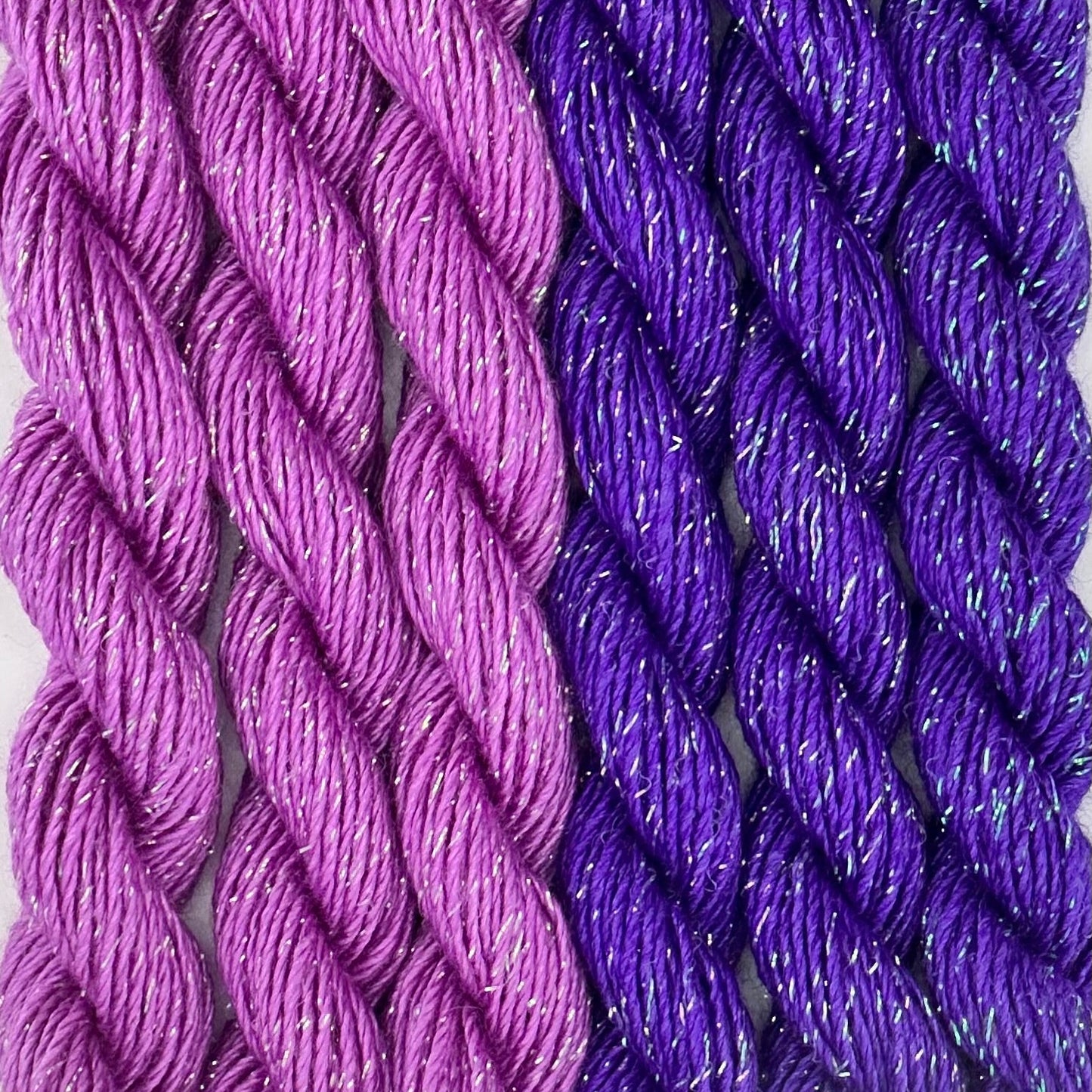 close-up of 6 skeins of Stardust Silk and Ivory needlepoint thread in two shades of purple including bacchus 651 and petunia 694, lying side by side.