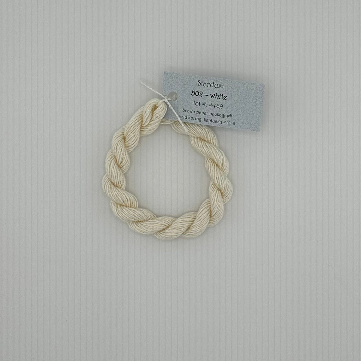 30 yard skein of Stardust Silk and Ivory needlepoint thread in white 502, with light blue label lying flat on white background