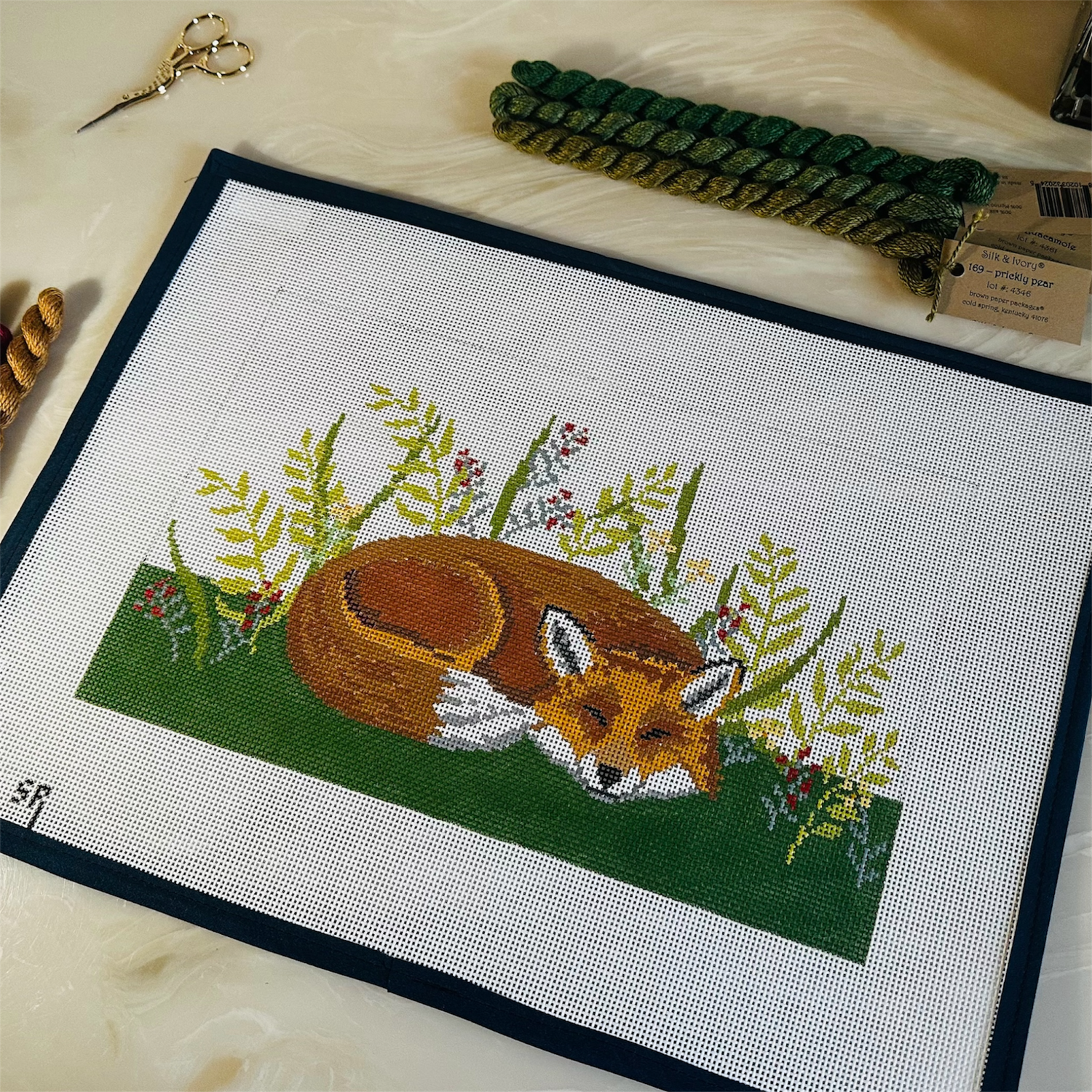 A susan roberts needlepoint canvas featuring a sleeping fox surrounded by green leaves and small flowers, bordered in dark blue. The canvas is laid on a light beige counter with green and brown silk and ivory threads and embroidery scissors.